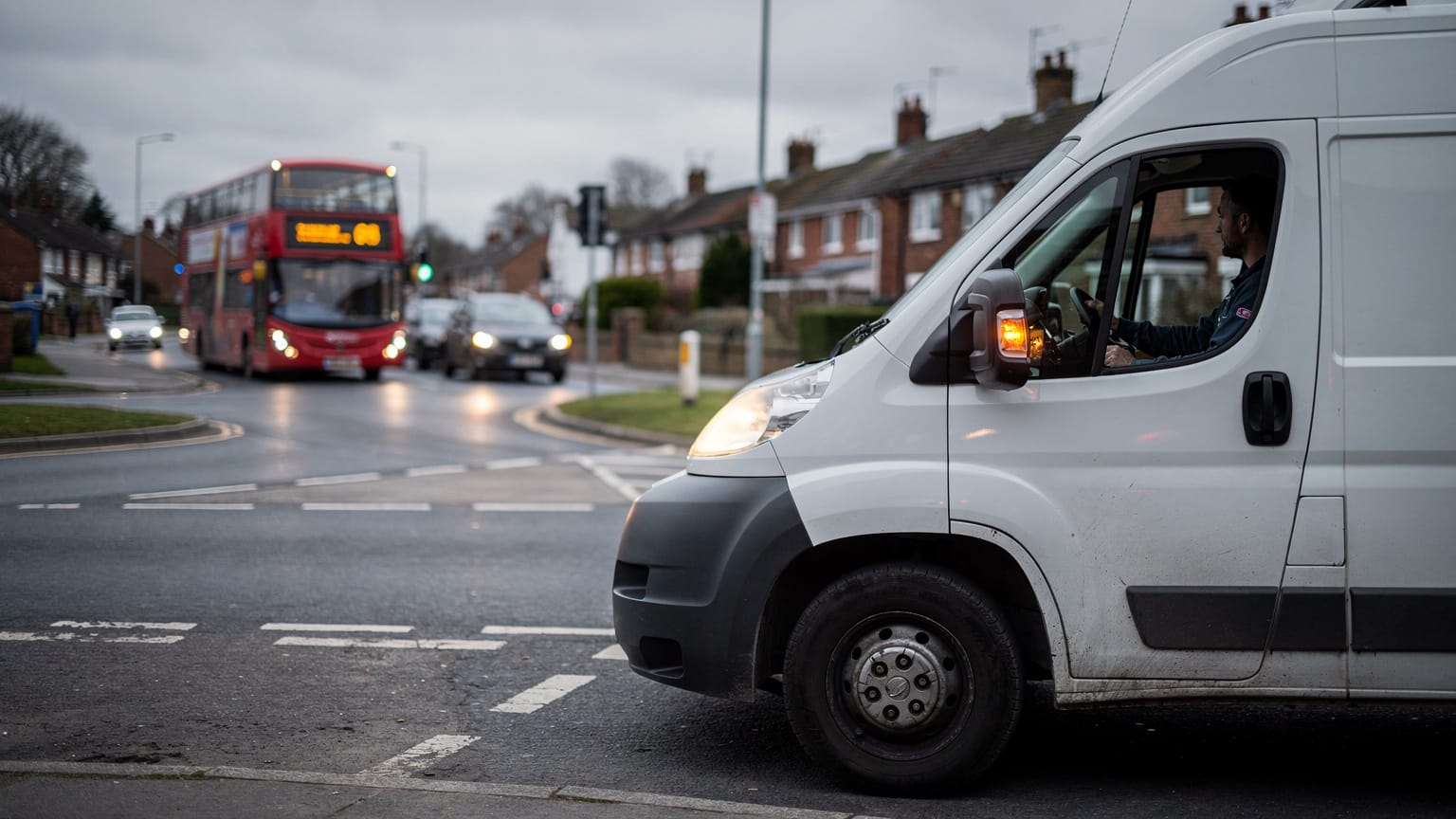 A white van waits at a suburban junction at dusk while a red double-decker bus and cars approach along a wet road lined with brick houses.